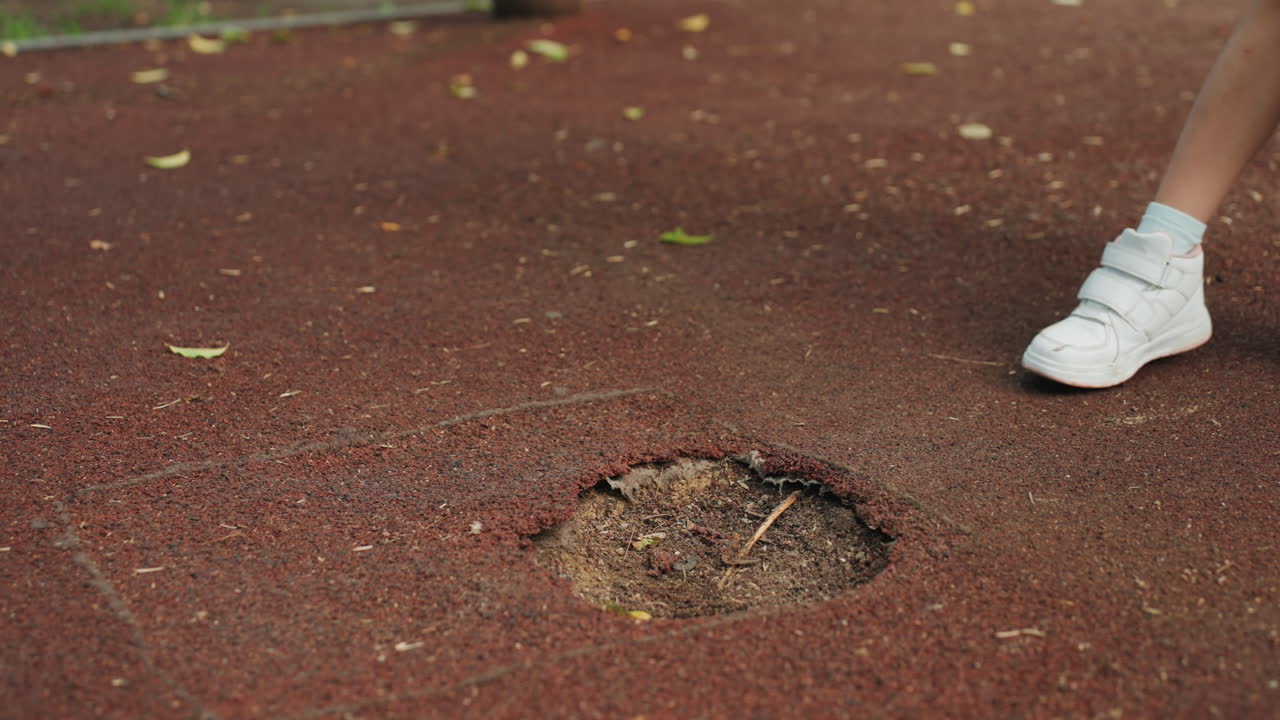 Dekat lubang di taman bermain, sepatu kets anak perempuan terlihat, ujung sepatu kets putih di samping lubang karet dangkal, daun berserakan dan kerikil di dalamnya, permukaan pengaman merah menunjukkan keausan, menyiratkan langkah ragu-ragu.