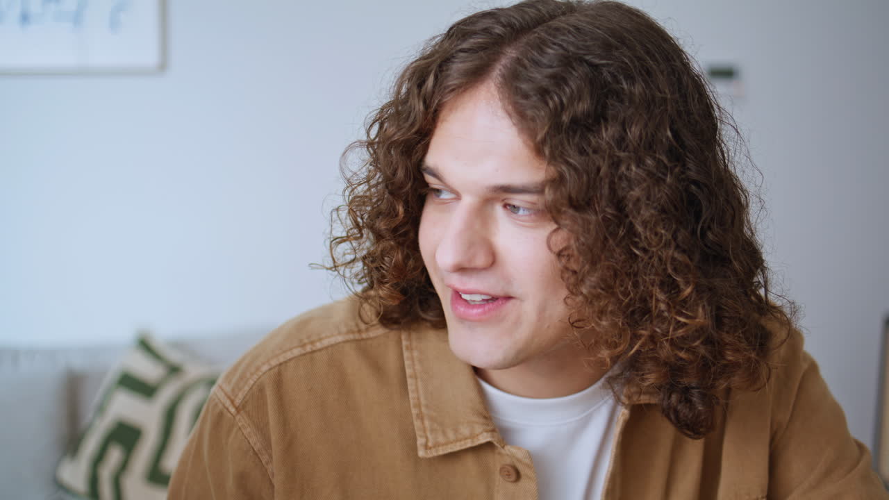 Guy sipping orange juice smiling to friend in apartment closeup. Curly-hair man
