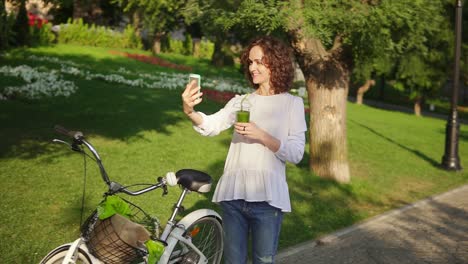 portrait of a young woman taking selfie photo with cell phone standing in the city park near her city bicycle with flowers in its