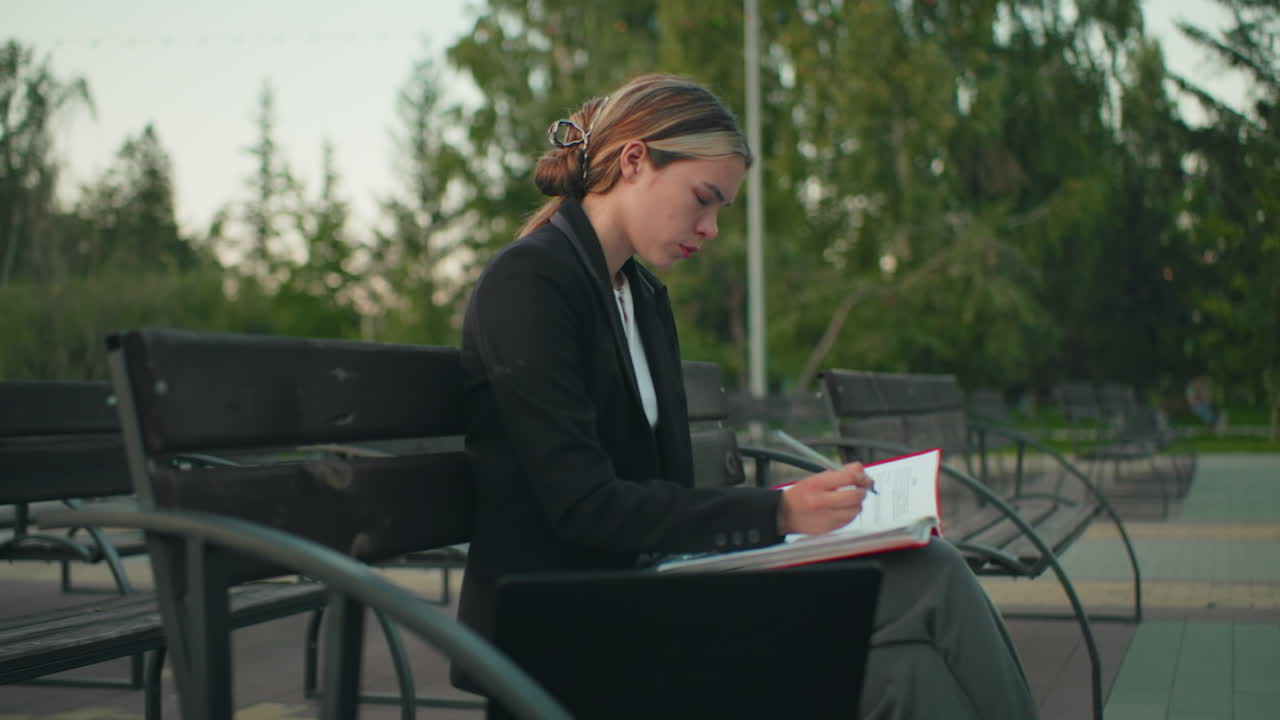 Student in professional attire seated alone outdoors focused on writing in red folder with pen, laptop beside her on bench in quiet park with trees and empty benches in background