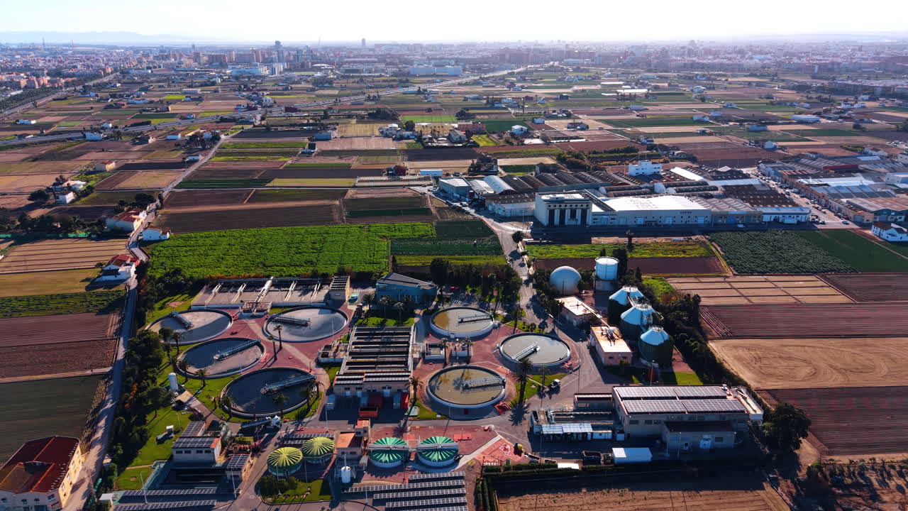 Wide aerial view of water treatment plant and agricultural fields. High angle view showing an industrial water processing facility surrounded by farmland and a city skyline in Spain
