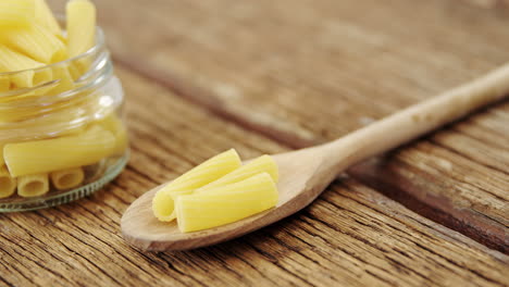 tube pasta  in one little glass jar and wooden spoon on wooden background