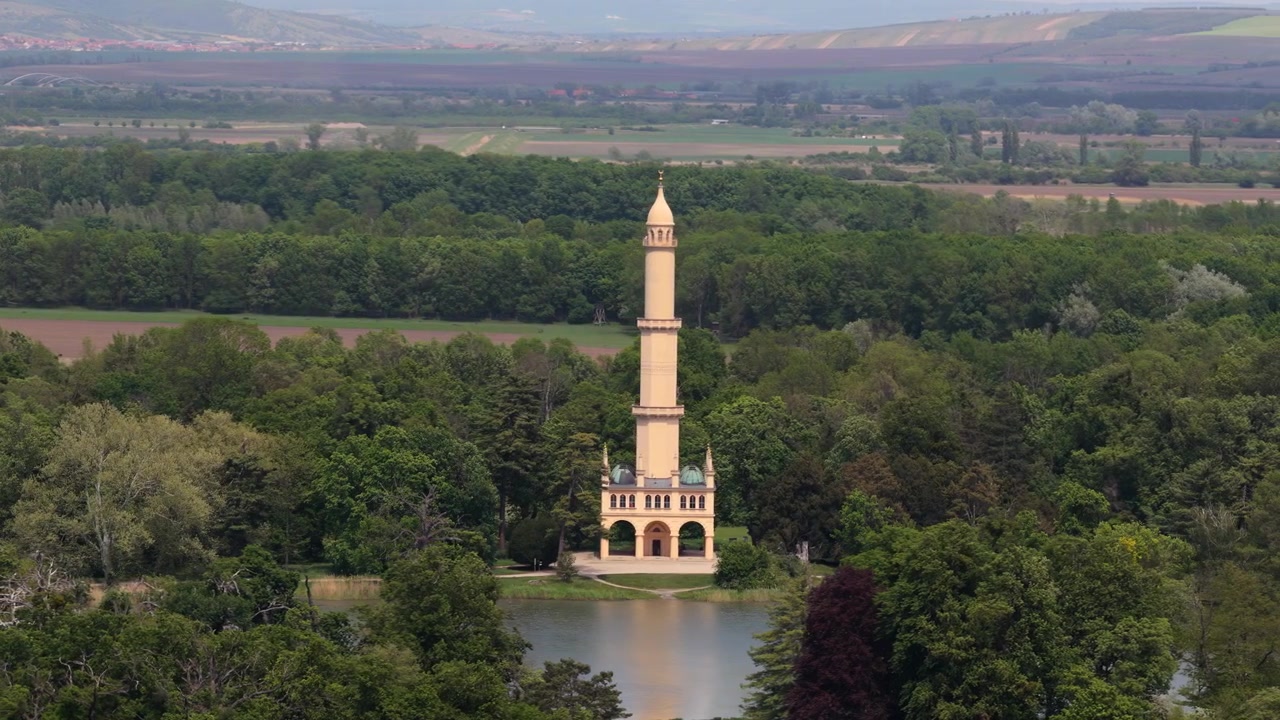 Dronekuva korkeasta maurilaisesta minareetista ja paviljongista puiston järven rannalla, jota ympäröi rehevä metsä, ja etäällä näkyy tasaisia viljelysmaan tasankoja, Moravia, Tšekin tasavalta.