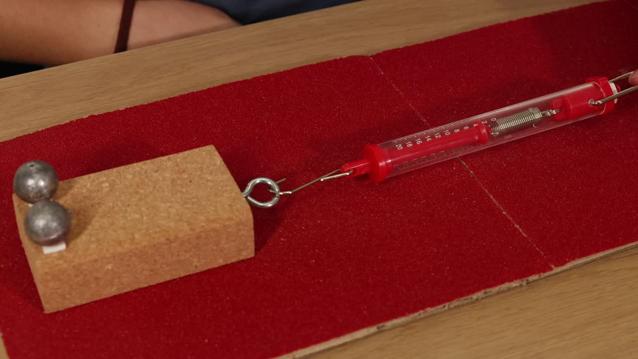Hands use a spring scale to measure frictional force on a block with weights on a red surface