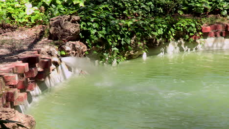 Pequeño-Lago-De-Agua-Verdosa,-Con-Una-Fuente-De-Agua-Corriente,-Un-Patito-Que-Empieza-A-Nadar