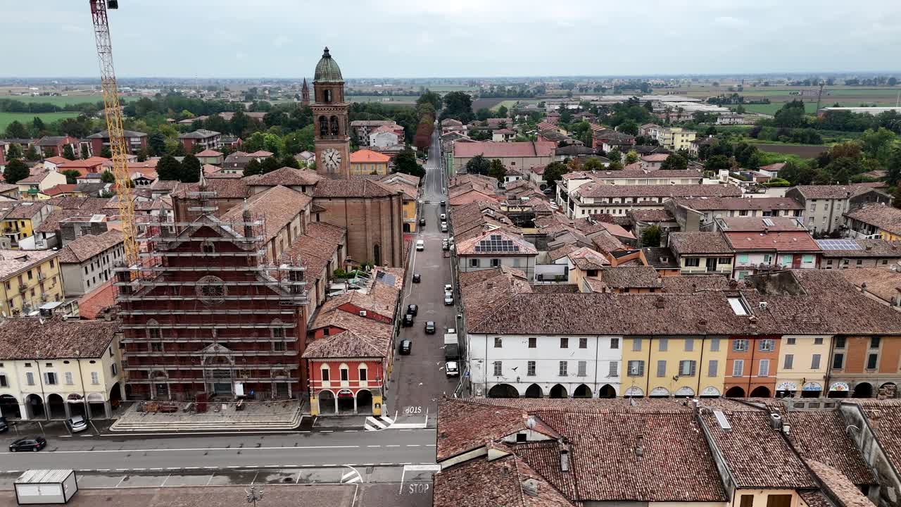 Aerial drone pan reveals the charming historic center of Cortemaggiore PC Italy featuring a prominent church undergoing renovation surrounded by tiled rooftops