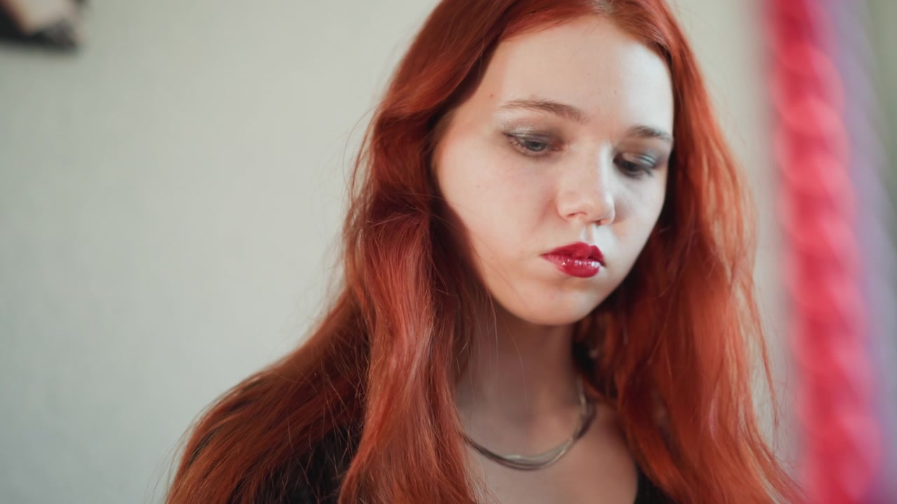 Primo piano di donna caucasica dai capelli rossi pensierosa, luce interna soffusa, trucco glitterato per le sopracciglia, collana d'argento a strati, lunghi capelli castano ramato, quieta malinconia, sottili movimenti della testa, atmosfera cinematografica intima