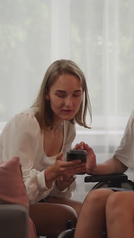 blonde woman shows photos from vacation to girl friend with disability in wheelchair. cheerful ladies look in smartphone and smile sharing happy memories