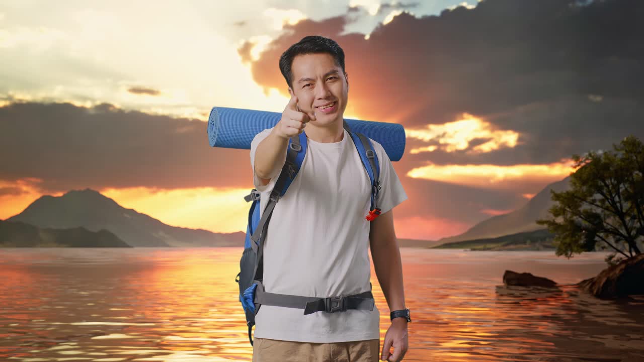 Asian Male Hiker With Mountaineering Backpack Smiling, Touching His Chest, And Pointing To Camera At A Lake