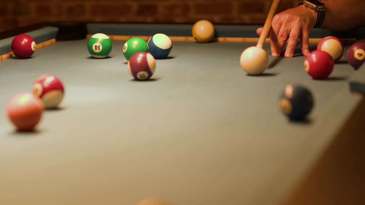 Hand strikes cue ball, scattering billiard balls across felt table under warm, low lighting