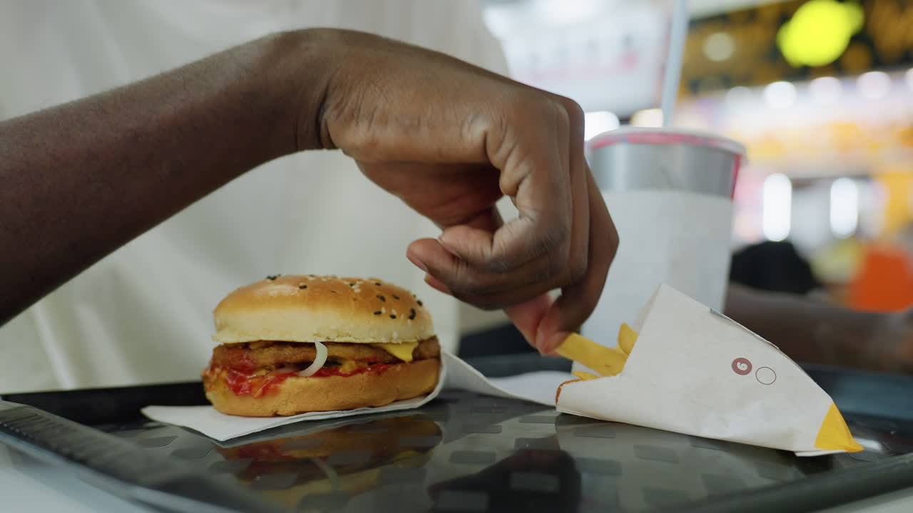 Close up hand reaching toward sesame bun burger with ketchup onion patty next to fries and cold drink on tray inside food court, bright modern interior with bokeh lights and visible human presence
