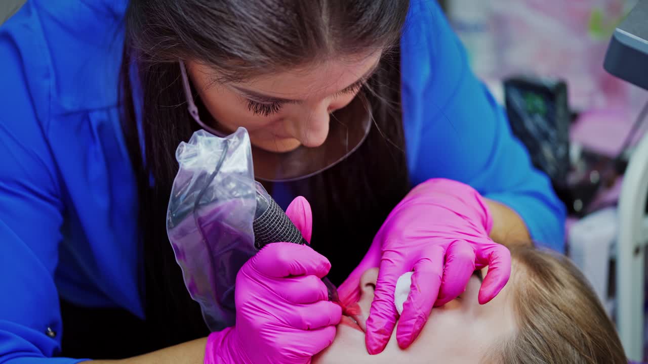 Lips tattooing in beauty salon. Professional cosmetologist in pink gloves doing permanent makeup on lips with a special instrument.