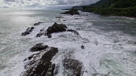 aerial dolly in over rugged shore and foamy sea waves near dense forest hillside in dominicalito beach, costa rica