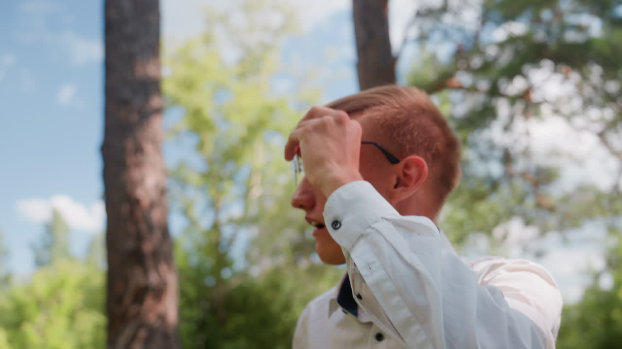 Portrait view of man in white shirt adjusting glasses while walking through forest path, carefully observing trees and nature around him under clear blue sky on bright summer day