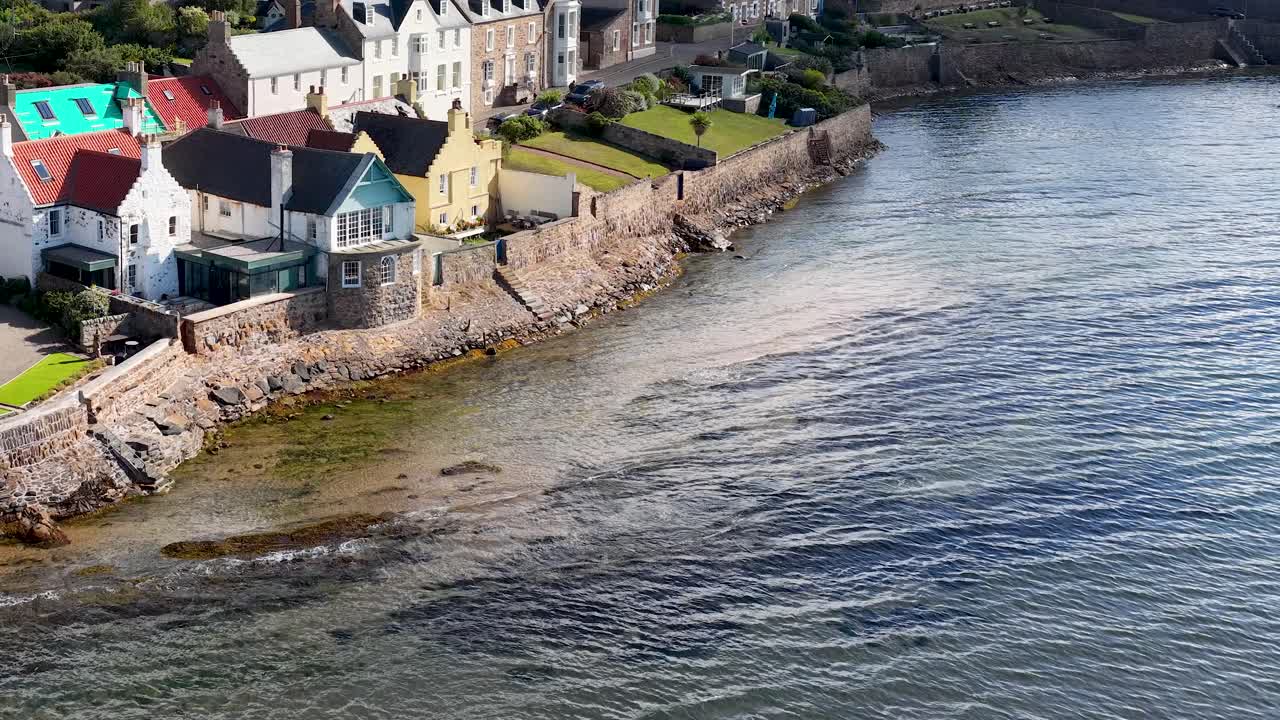 Drone camera smoothly pans above historic seaside village, colorful rooftops, stone seawall, and calm water