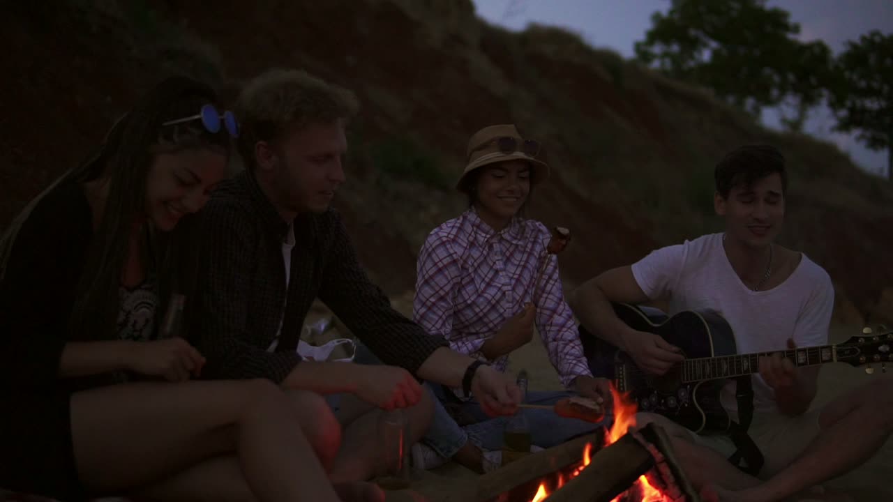 grupo de jóvenes y alegres sentados junto al fuego en la playa por la noche, asando salchichas y tocando la guitarra