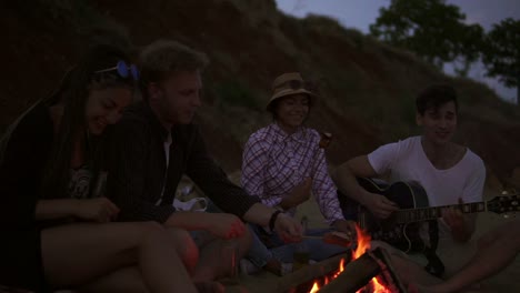 group of young and cheerful people sitting by the fire on the beach in the evening, grilling sausages and playing guitar