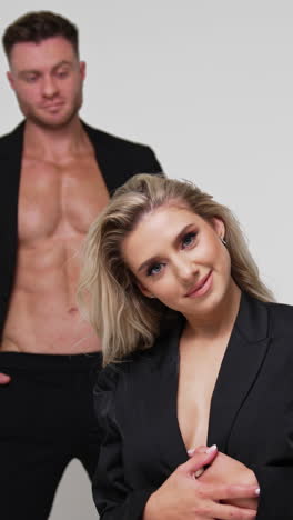 beautiful long-haired blonde woman stands on her knees waving her hair. man in black suit stands at backdrop. white background. vertical video