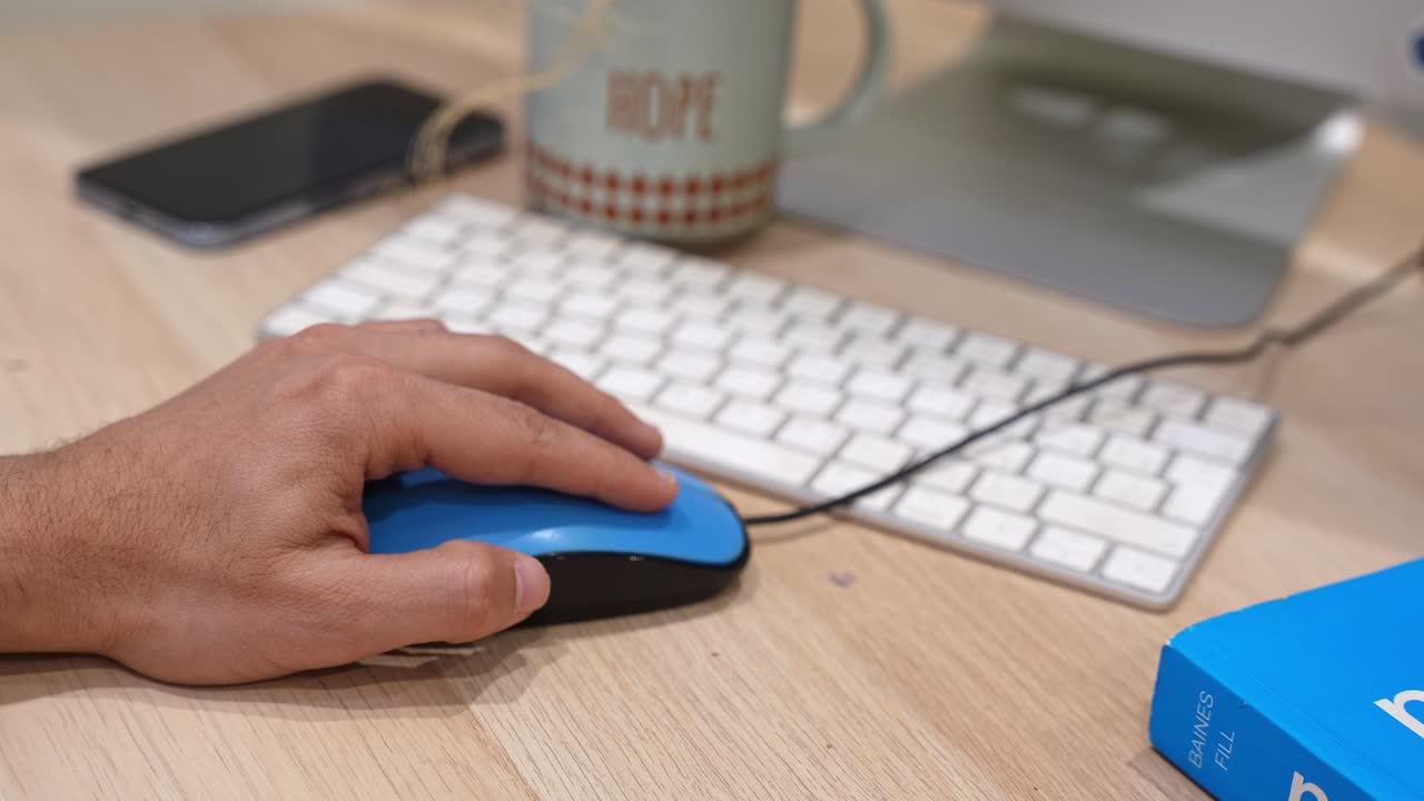 Close-up of a hand using a blue computer mouse beside a white keyboard, smartphone, and coffee mug on a wooden desk in a modern office workspace