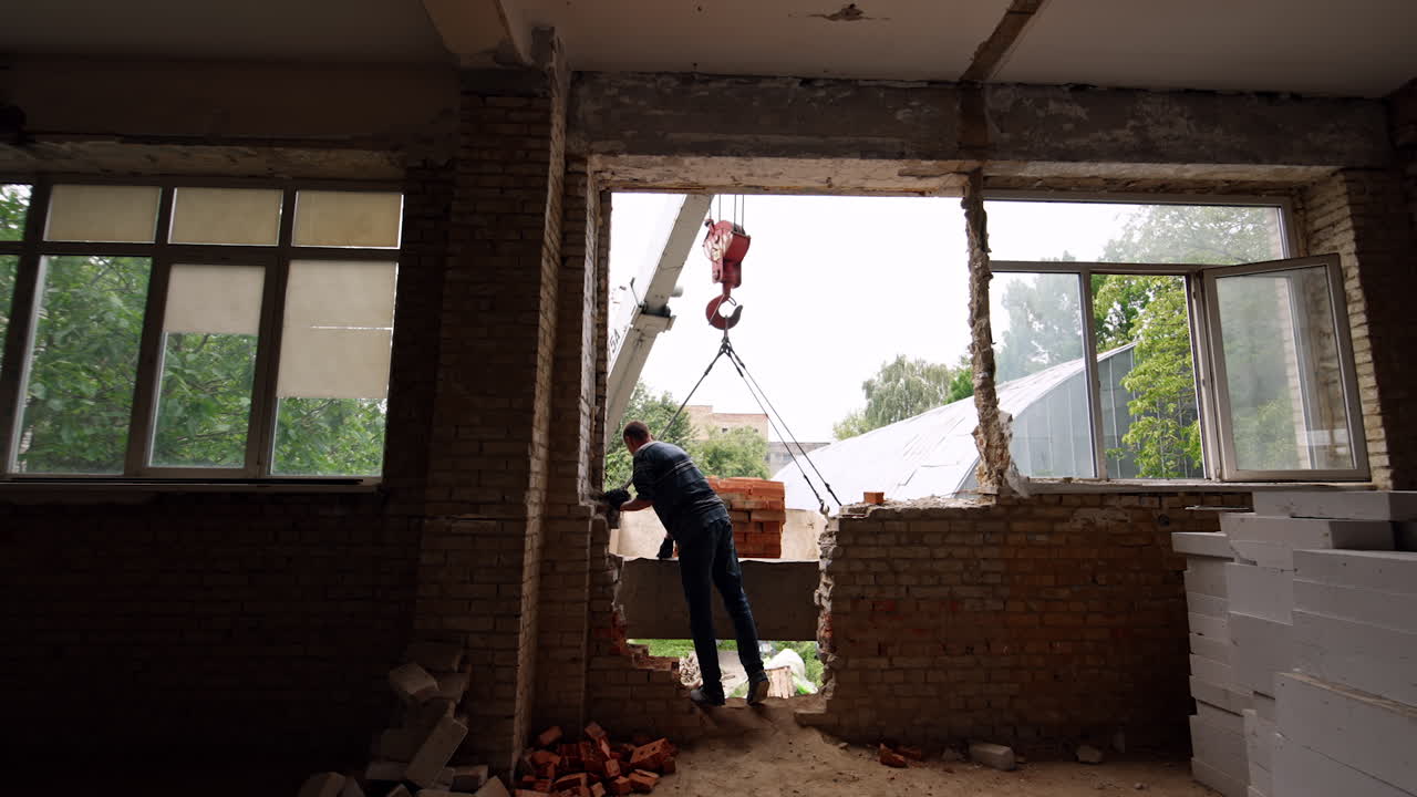 Approaching a man standing at the broken wall inside the building. Builder is watching and controlling the excavator lifting the bricks.