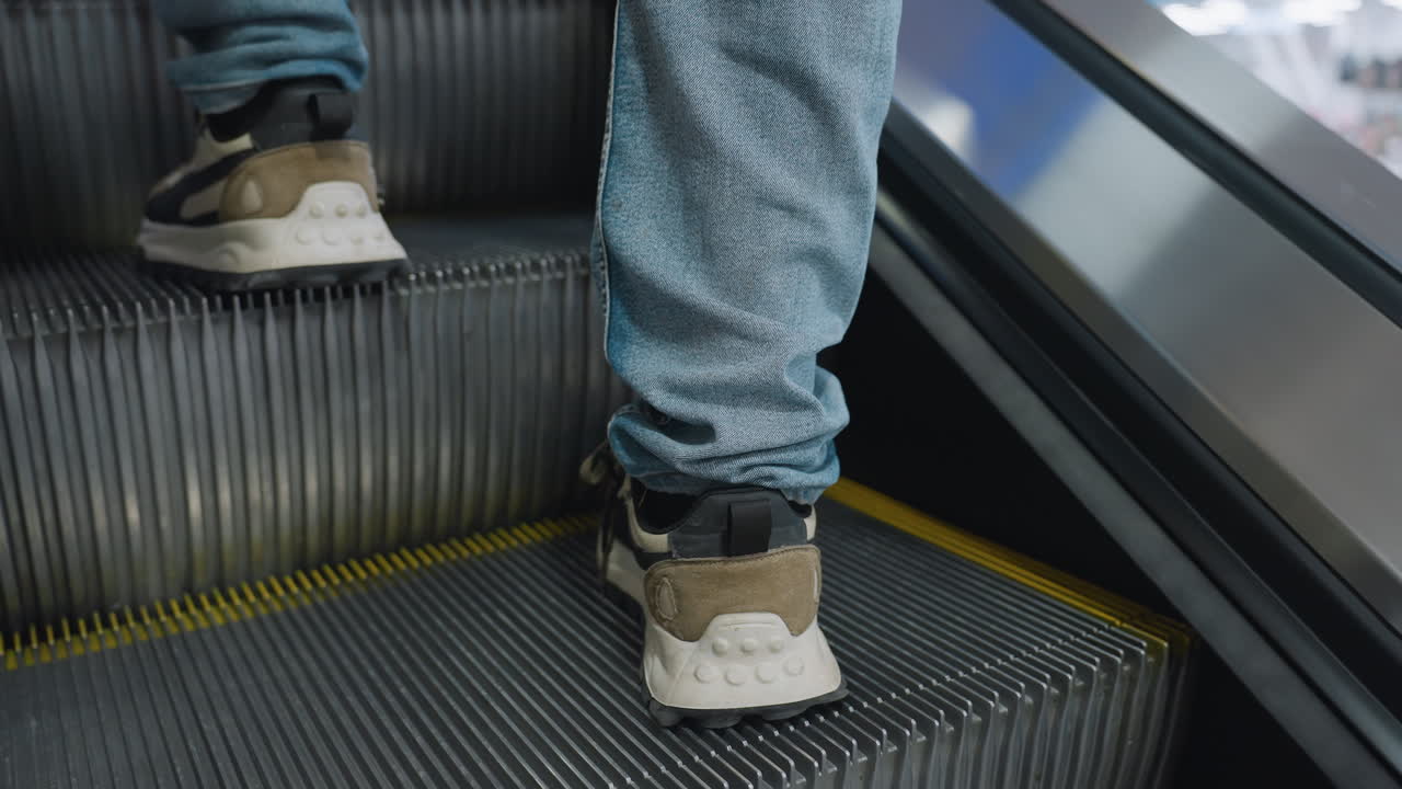 Nærbillede af person bagfra iført jeans og sneakers, der går op ad en rullende rulletrappe med synlige metaltrin, gule sikkerhedslinjer og slørede refleksioner af et lyst centerinteriør gennem glaspaneler