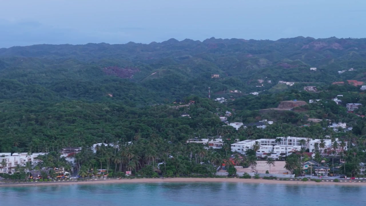 Ripresa aerea con drone che panoramica lungo la costa di Las Terrenas, rivelando hotel sulla spiaggia, lussureggianti colline verdi e una prominente punta sabbiosa durante il crepuscolo