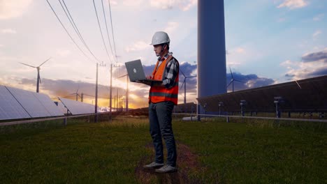 full body side view of asian male engineer with safety helmet working on a laptop while standing with solar panel and wind turbines