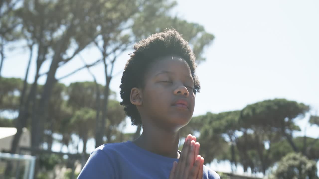 Adolescente afroamericano che prega, con le mani giunte in maglietta blu tra i pini mentre la telecamera si allontana.