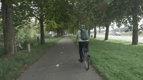 Slow-motion-footage-behind-a-bicyclist-wearing-a-green-backpack-riding-on-a-road-through-the-dutch-countryside