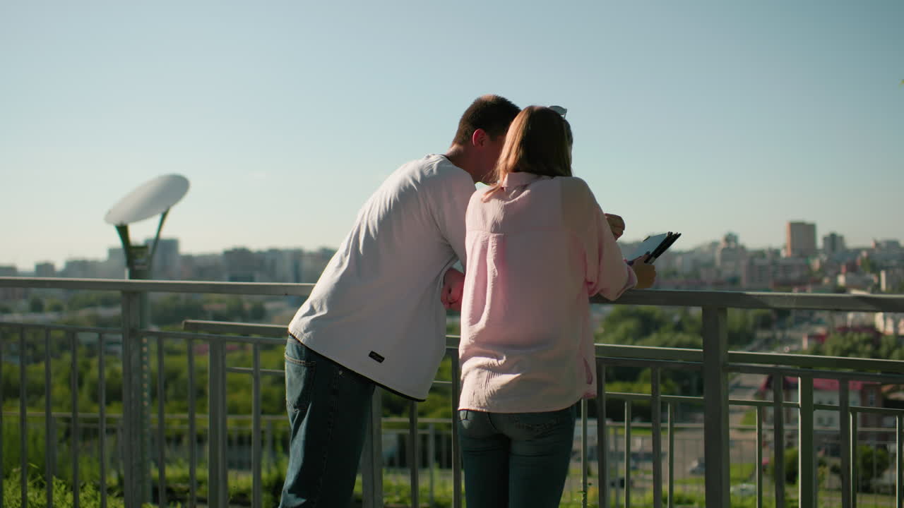 소년과 소녀가 철의 레일링 위에 기대어 상호 작용하는 측면 뷰 소년은 태블릿에 무언가를 보여줍니다. 배경은 초록색과 밝은 빛으로 흐릿한 도시 풍경을 특징으로합니다.