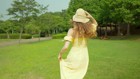 Playful-woman-in-yellow-dress-running-in-park-and-turn-back-looking-at-camera,-slow-motion