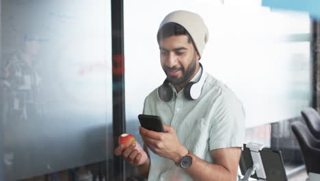 Un-Joven-Hombre-De-Negocios-Asiático-Con-Barba-Sonríe-Mientras-Usa-Su-Teléfono-Inteligente,-Sosteniendo-Una-Manzana