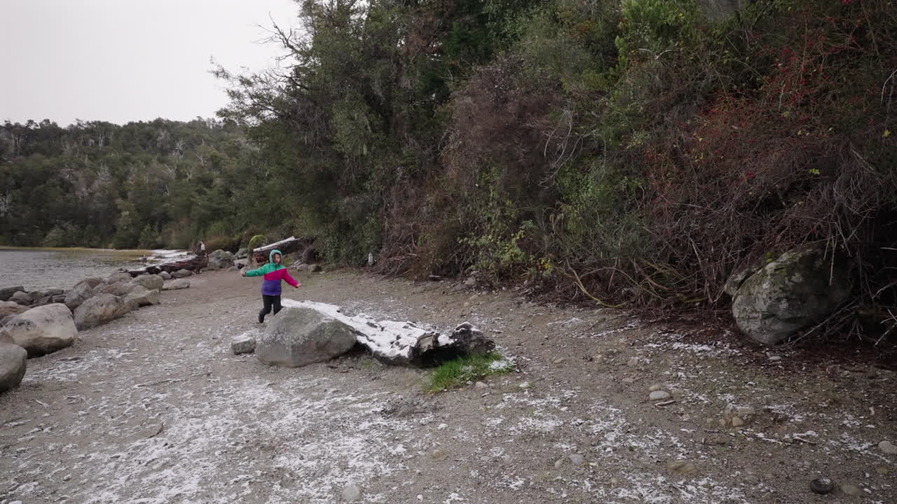 Boy dressed in winter clothing almost loses balance on an icy rock near a snow-covered lakeshore, catching himself mid-slip as strong wind and light snowfall sweep across the cold scene