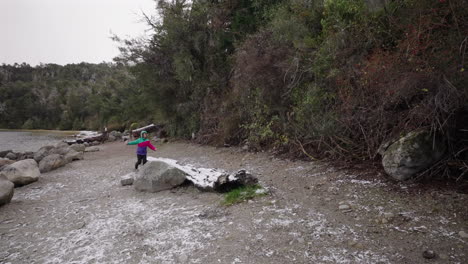 boy dressed in winter clothing almost loses balance on an icy rock near a snow-covered lakeshore, catching himself mid-slip as strong wind and light snowfall sweep across the cold scene