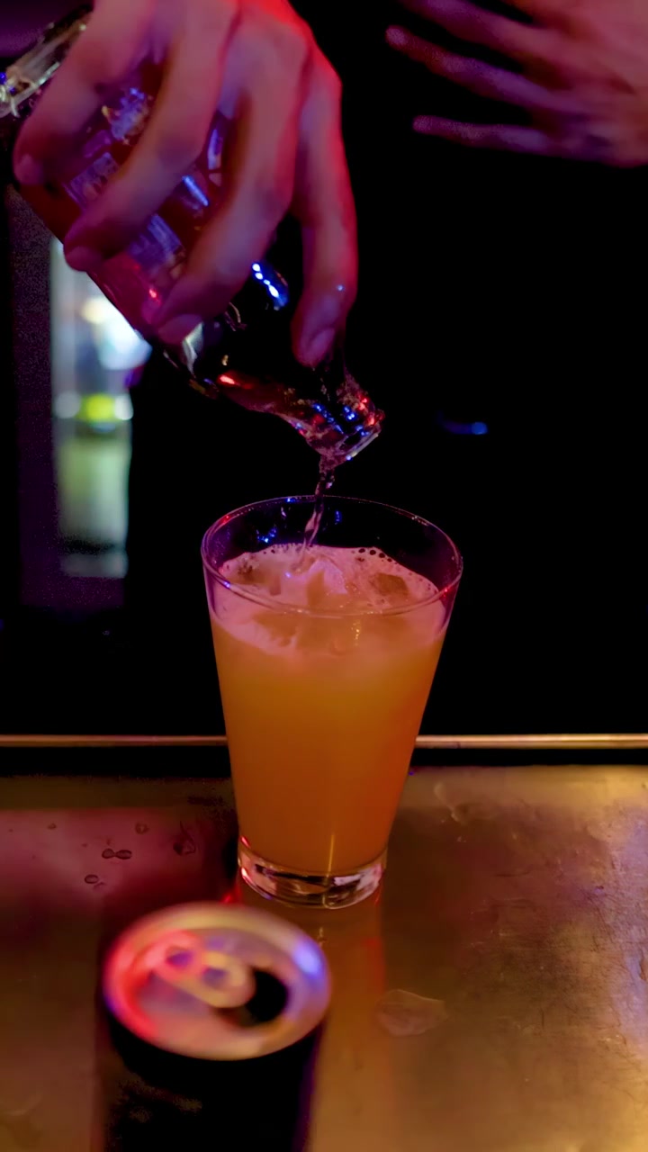 Un barista versa soda trasparente in un cocktail arancione con ghiaccio sotto un'illuminazione colorata del bar, vista ravvicinata.
