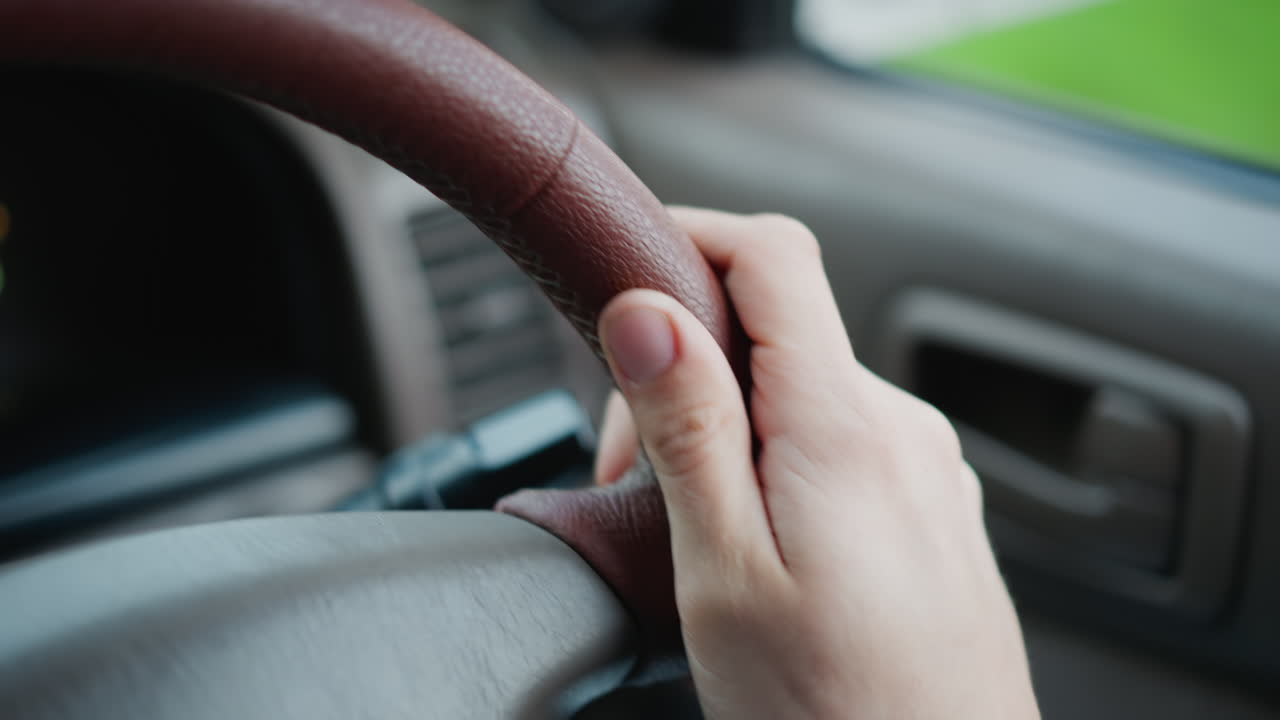 Primer plano de la mano derecha en el volante, conducción suburbana diurna con luz solar, agarre firme en el volante de cuero al pasar por una carretera verde, luces del salpicadero tenues, luz natural nítida y enfoque superficial, estable