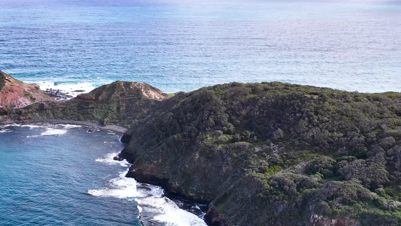 Drone glides above rocky coastline, lush headland, and blue ocean under natural daylight