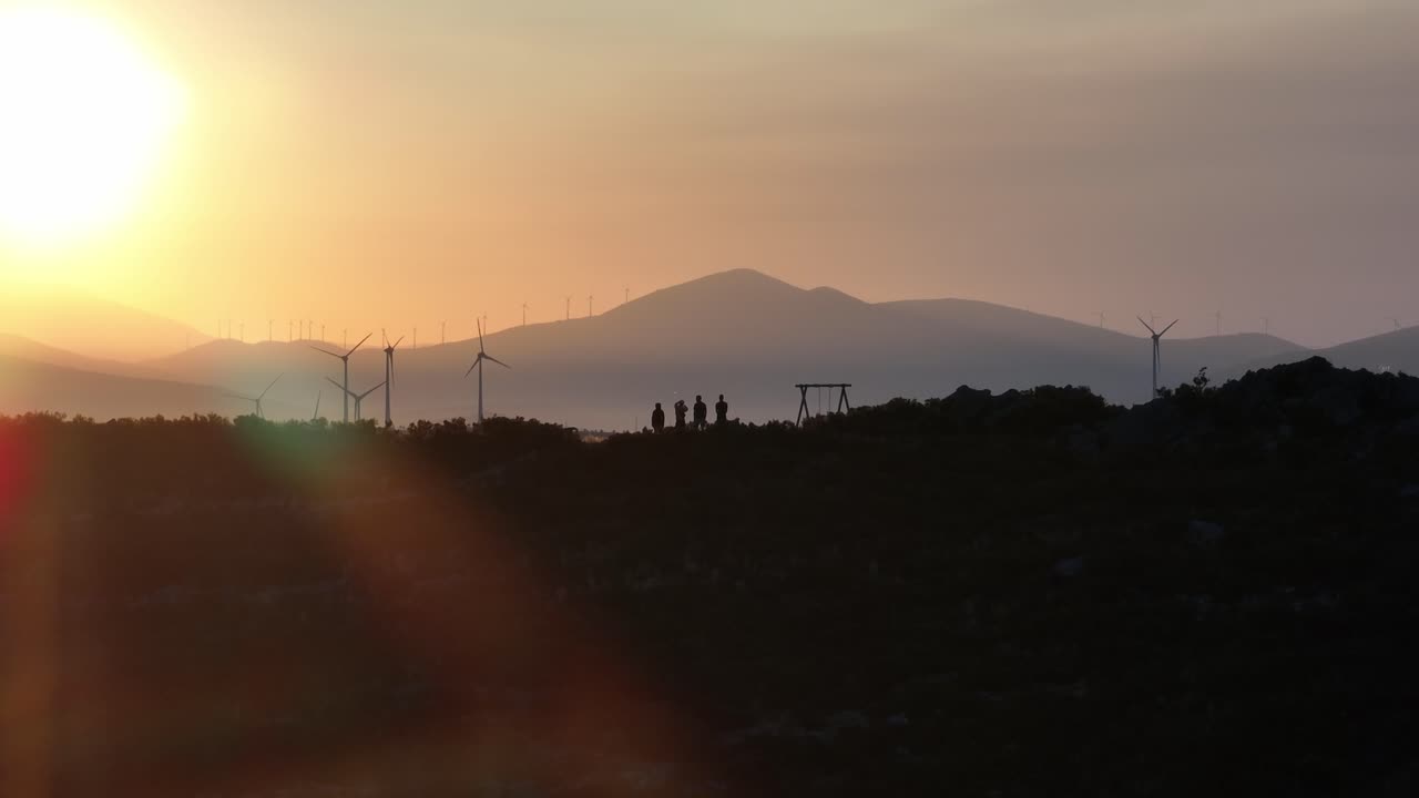 4 People standing on a hill in front of wind turbines. Silhouettes shortly after sunrise.