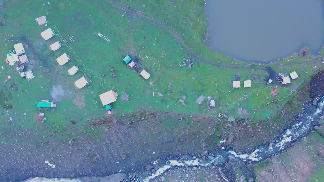 Top-down view of a riverside lake with scattered summer tents and flowing stream in Kashmir