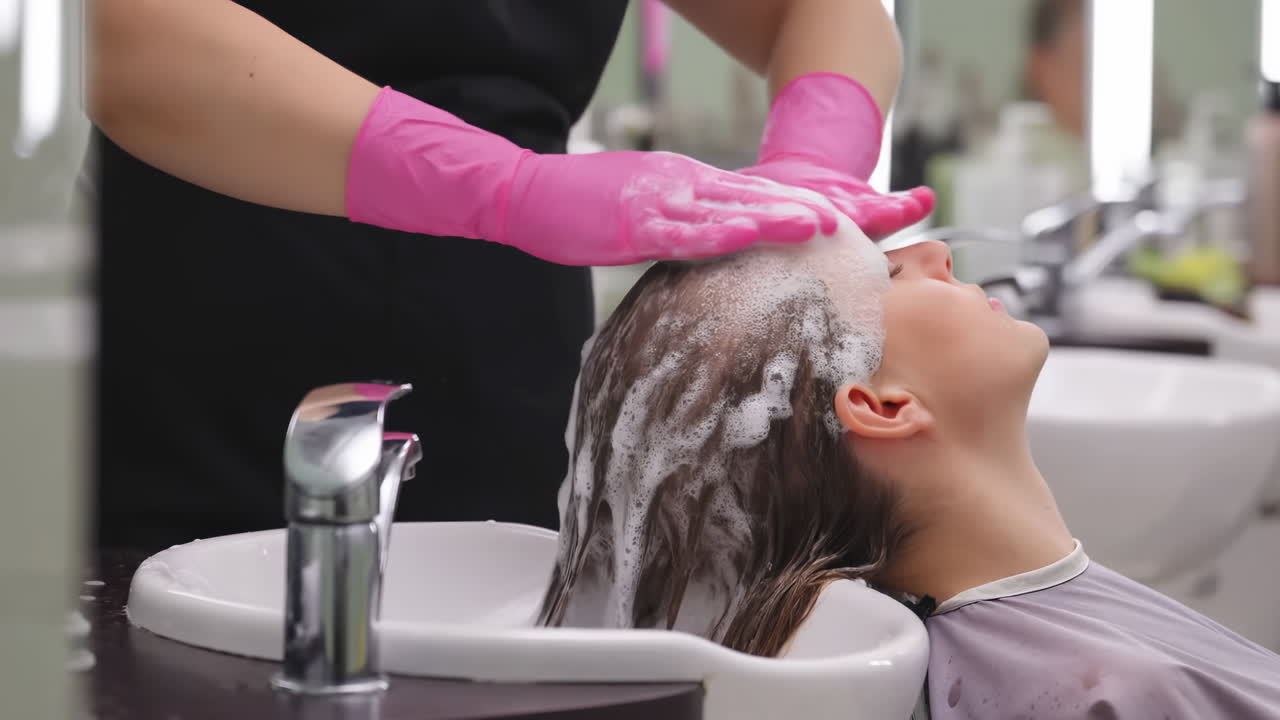 Donna che fa lo shampoo in salone, stilista con guanti rosa acceso che massaggia delicatamente il cuoio capelluto sopra una vasca bianca.
