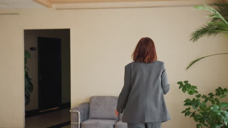 현대적인 사무실에서 소파로부터 멀어지는 여성 사업가, 음료를 들고 있고, 화분들이 근처에 있음, 캐주얼한 휴식 시간, 편안한 자세, 전문적인 복장, 사무실 라운지 공간