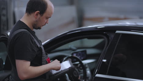 Portrait-A-mechanic-in-a-car-service-close-up-holds-a-tablet-in-his-hands-and-presses-on-the-screen-near-the-car