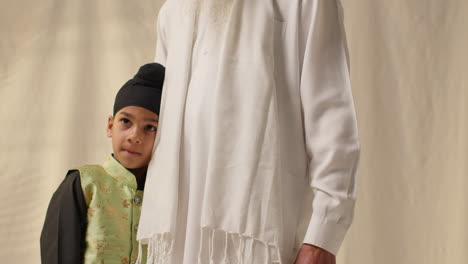 Studio-Shot-Of-Young-Sikh-Boy-With-Top-Knot-Turban-Hugging-Grandfather-Against-Plain-Background