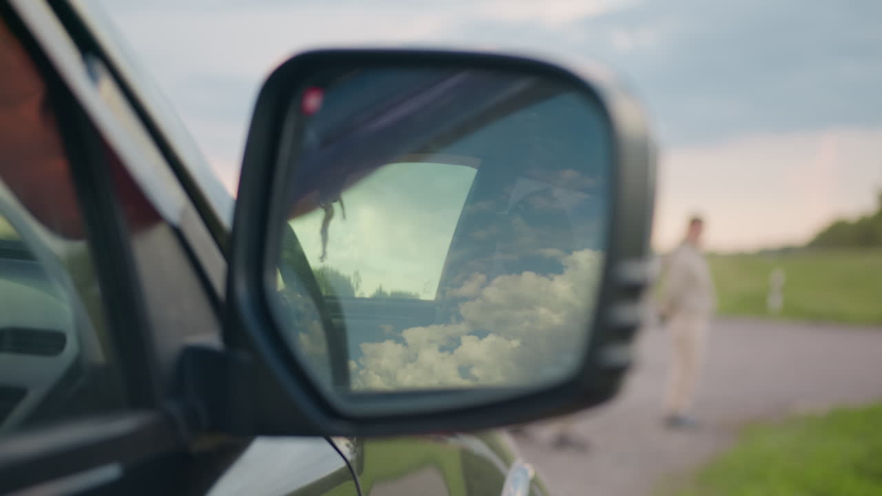 Close up of car side mirror reflecting bright cloudy sky while people in beige outfits stand in soft focus near grassy roadside, evoking calm atmosphere with contrast of nature and travel elements