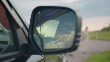 close up of car side mirror reflecting bright cloudy sky while people in beige outfits stand in soft focus near grassy roadside, evoking calm atmosphere with contrast of nature and travel elements