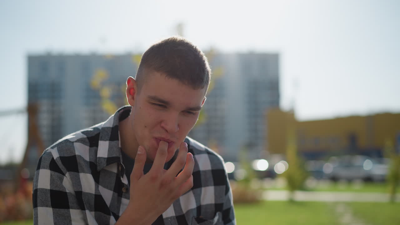 young boy seated outdoors during sunny day lifts hand to mouth and gently licks fingers while looking up with calm expression, background shows soft blur of buildings, trees, and blue sky
