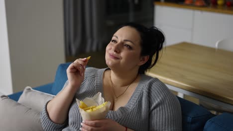 Plus-Size-Frau-Isst-Pommes-Frites-Mit-Großer-Freude-Auf-Einer-Couch-Sitzend