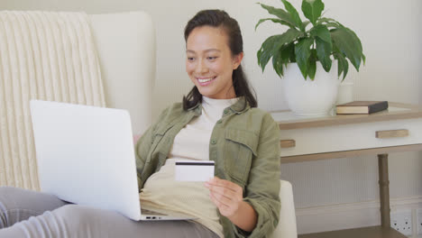 happy asian woman using laptop and holding credit card in bedroom, in slow motion