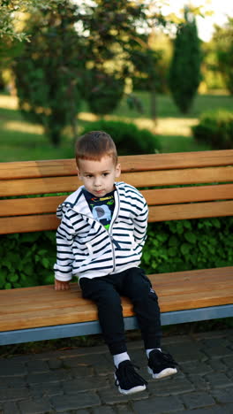 lovely kid sits down on the bench in the park. toddler boy sits to have rest during the walk. vertical video.