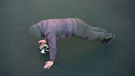 athlete performing push-ups alternating hands on soccer ball demonstrating body control on outdoor sports ground showcasing intense training and agility against plain green backdrop